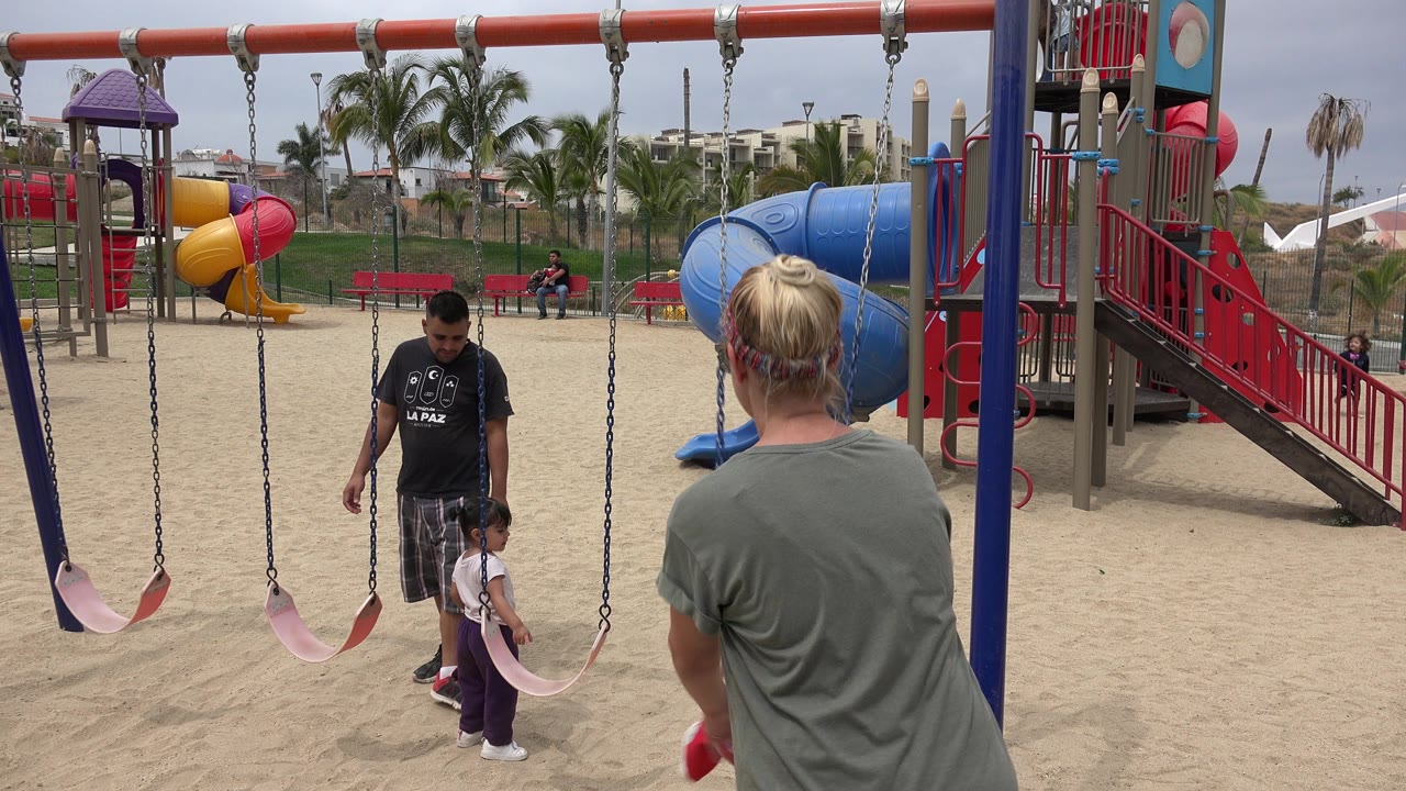 In San Jose Del Cabo, the man and small child stand closer to the swing seats while the blonde adult remains in the right foreground facing the playground.