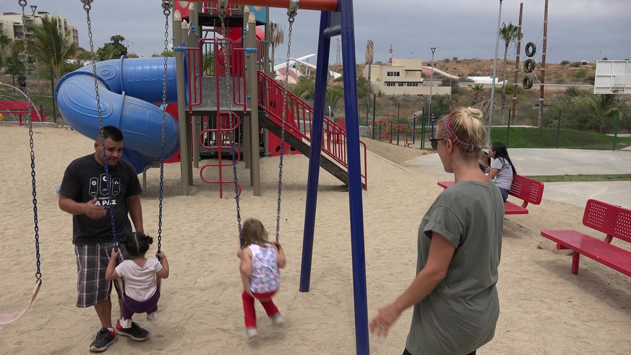 Adults and children at a sandy playground with swings and a blue slide.