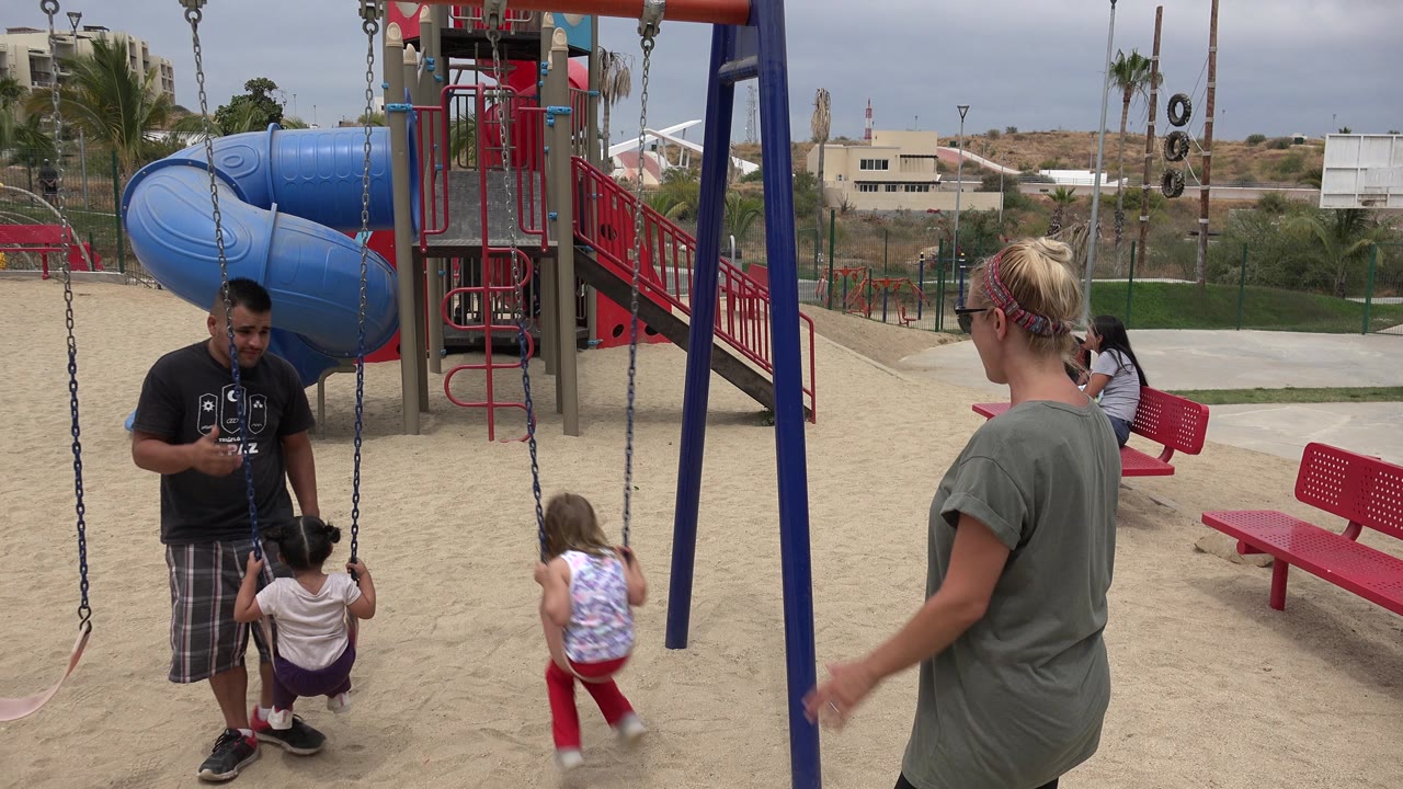 A girl in red pants moves toward the swing area as a woman in a gray shirt stands at right and a man watches another child on a swing in San Jose del Cabo.