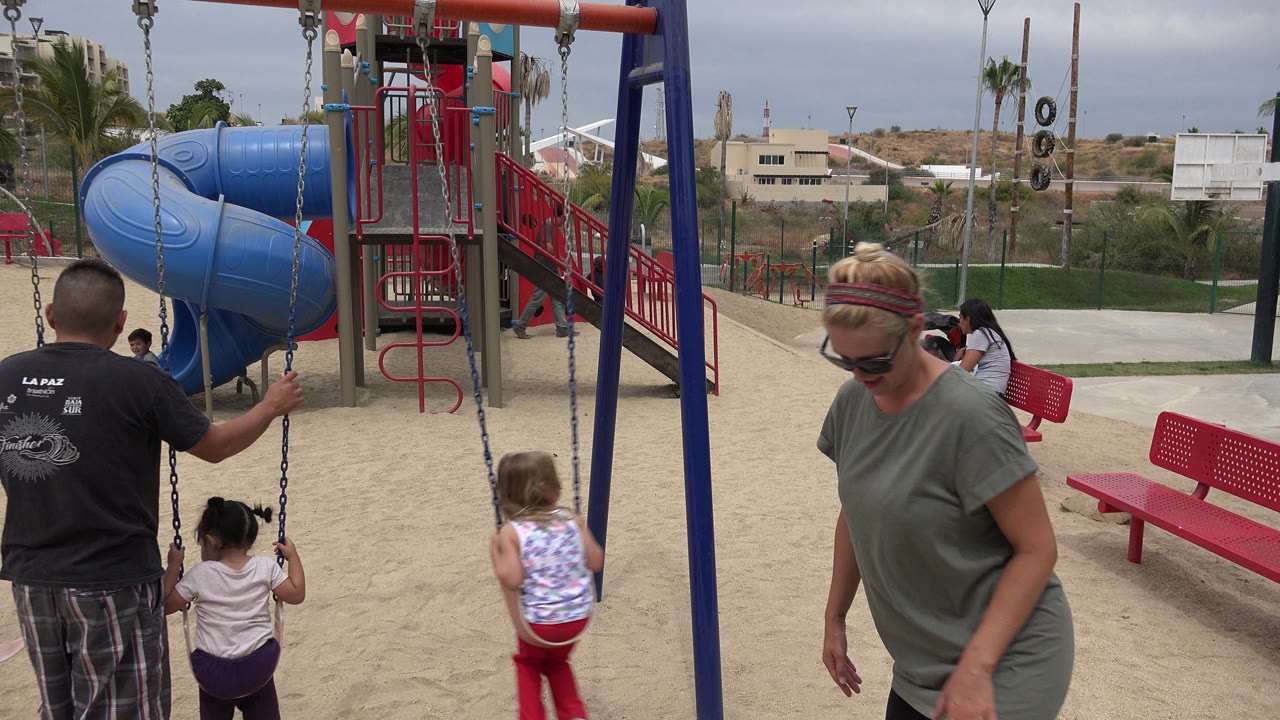 At a San Jose del Cabo playground, an adult man stands near swing chains as a young child sits on a swing and a second child approaches the sandy play area.