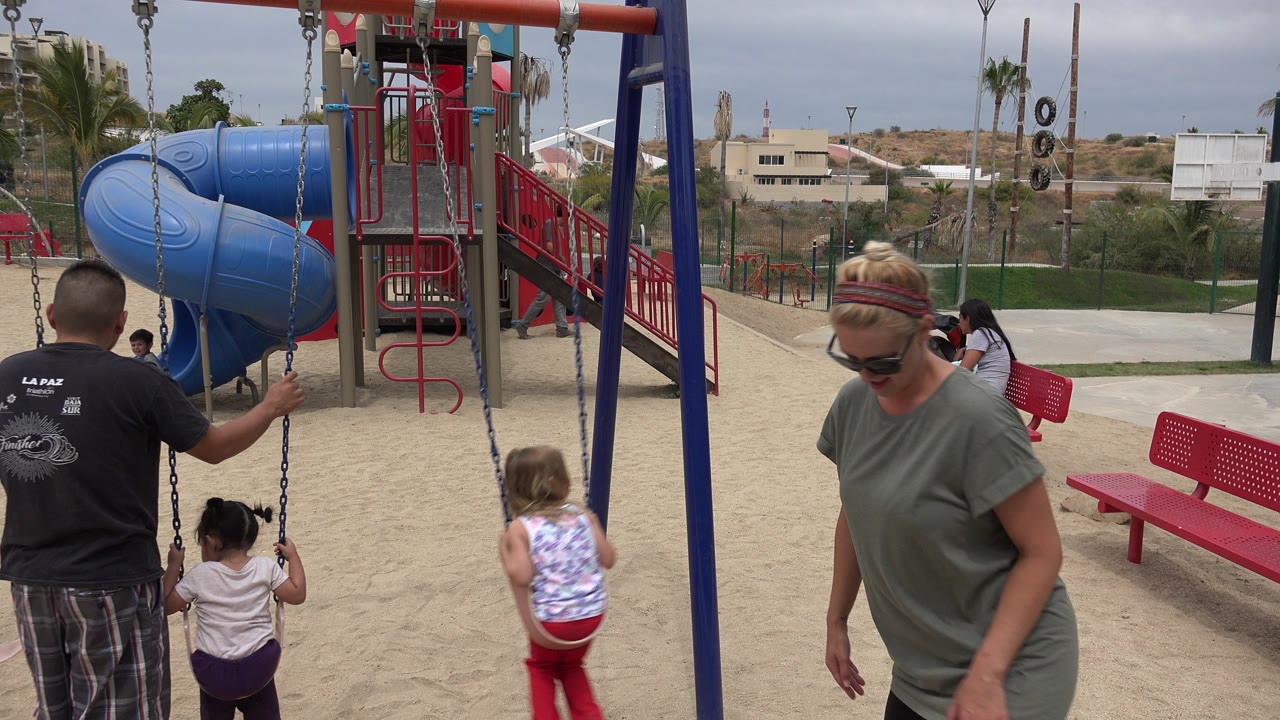 A blonde woman wearing sunglasses passes close to camera in San Jose del Cabo as children continue using the swings near the red and blue playground equipment.