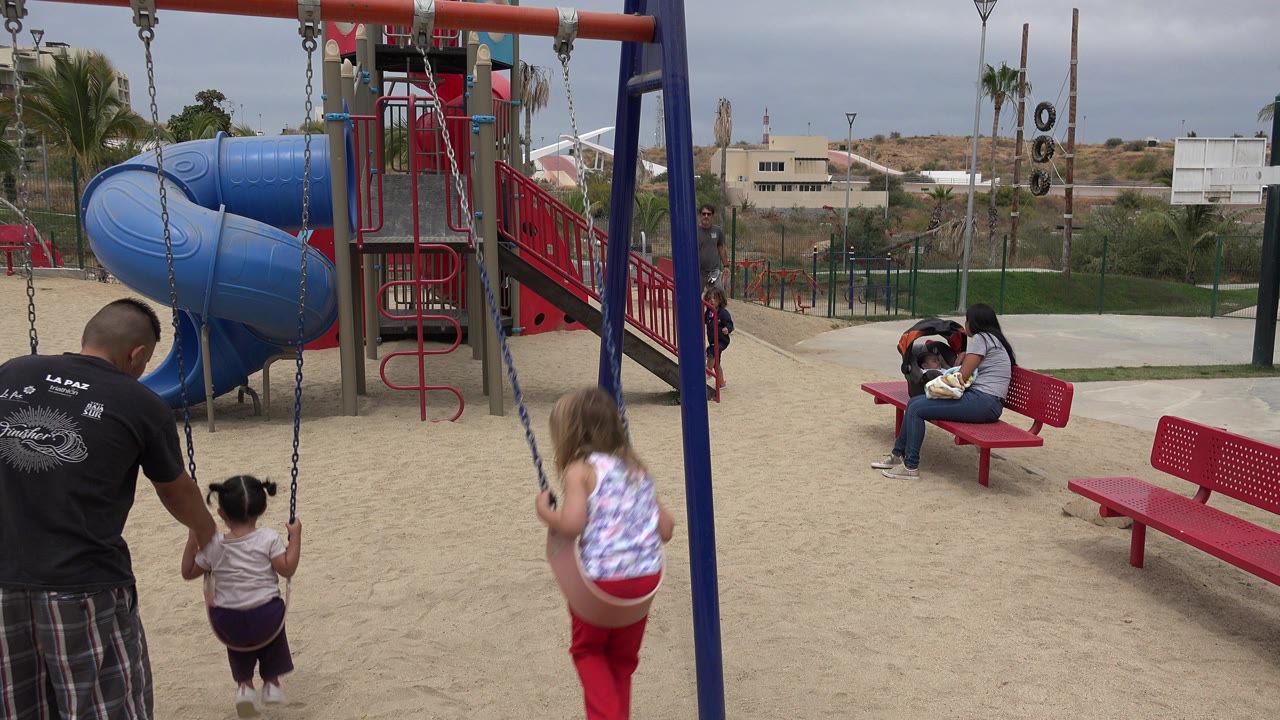Girl in red pants on a swing beside an adult and smaller child at a playground.
