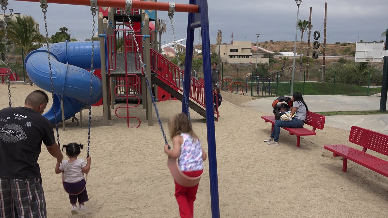 At 0:01 in San Jose del Cabo, the girl in red pants is higher on the right swing as an adult stands behind a smaller child, with the blue tube slide and red railings filling the background.