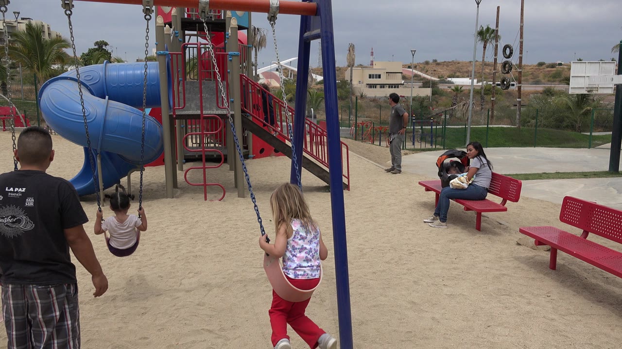 Father near two girls on playground swings beside a blue slide.