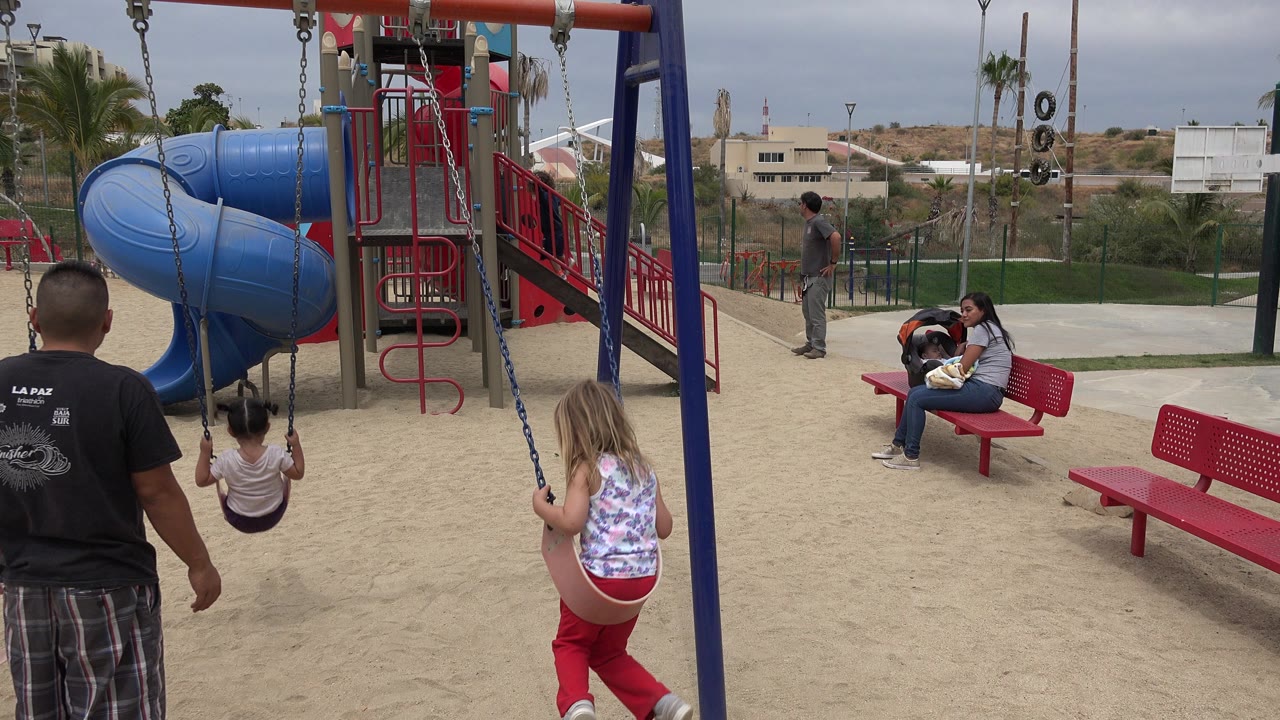 At 0:01 in San Jose del Cabo, Mexico, the father faces the swings as one girl sits forward and another girl swings nearby, with the blue slide and red railings filling the play area.