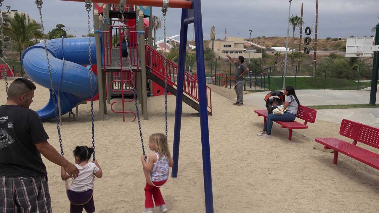 At 0:01 in San Jose del Cabo, the father stands closer to the center swing while one girl sits facing away and another girl stands by the adjacent swing chain.
