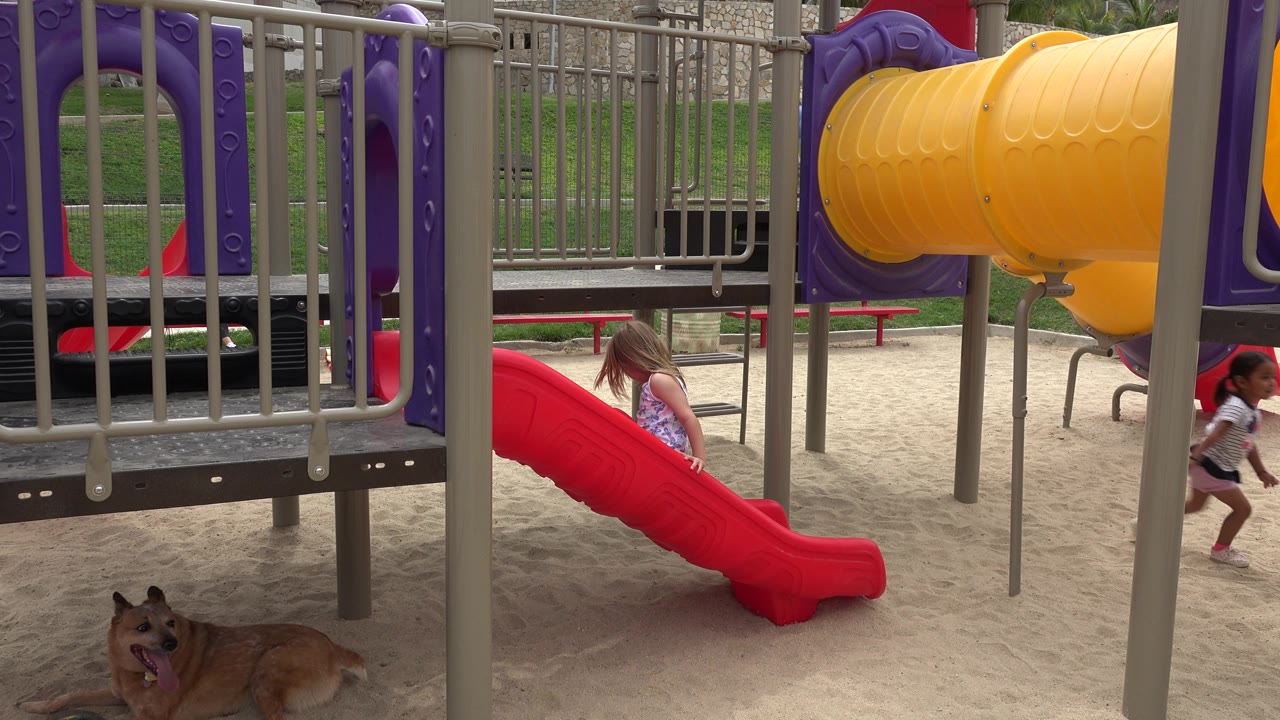 Girl stands near the red slide as another child runs at the right edge of the sandy playground.