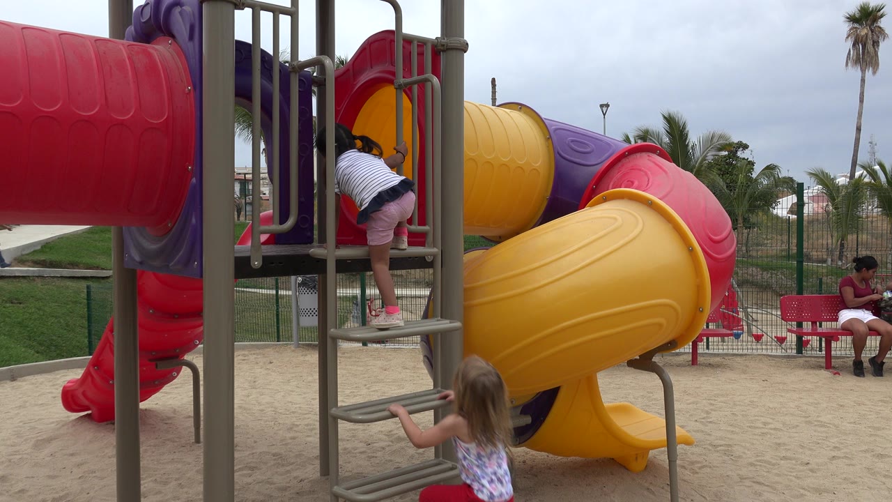 Two girls climbing a playground ladder beside colorful tube slides.