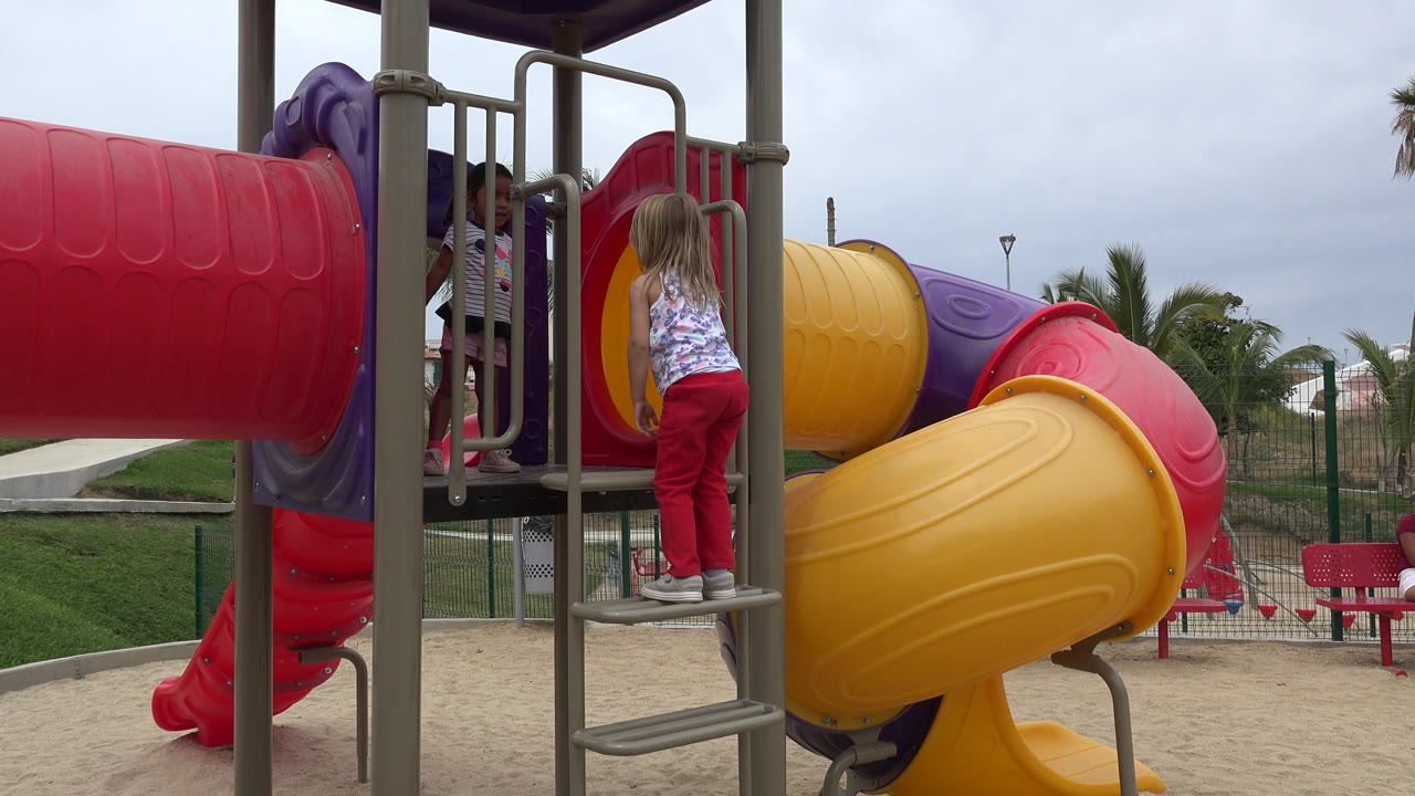 A blonde girl in red pants stands on a metal playground ladder beside red, yellow, and purple tube slides in San Jose Del Cabo, Mexico.