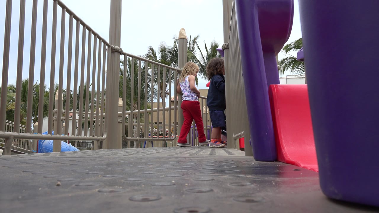 Two children stand on a playground platform in San Jose Del Cabo, Mexico, with tan railings, a red slide, a purple support, grey flooring, and palm trees behind them.