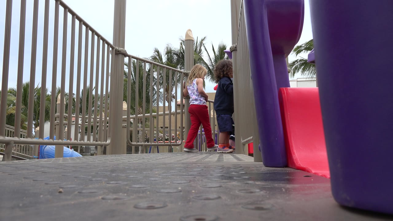 The children stay near the tan playground railings in San Jose Del Cabo, Mexico, while the red slide and purple support dominate the right side of the frame.