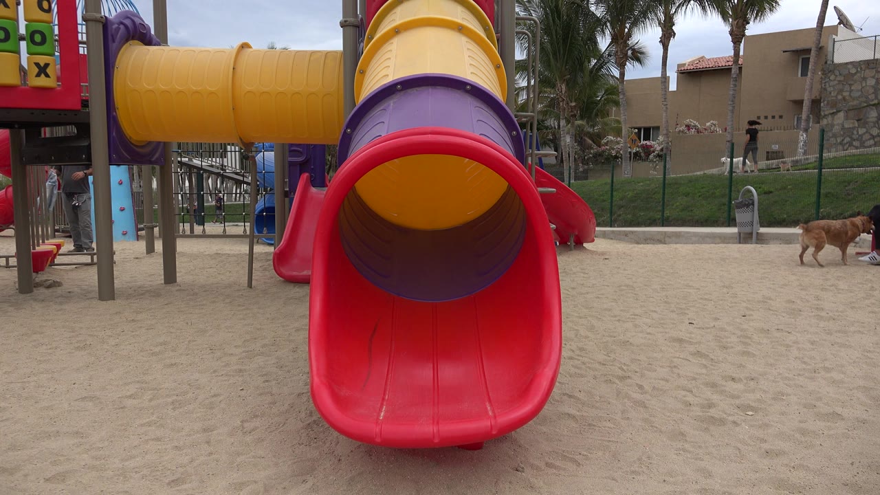 Red playground slide opening on sandy ground with palms and buildings behind it.