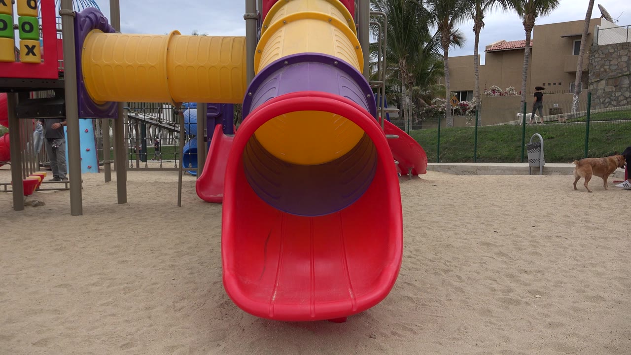Centered playground slide in San Jose del Cabo with a dog moving across the sandy area at right and distant people near the fence and play equipment.
