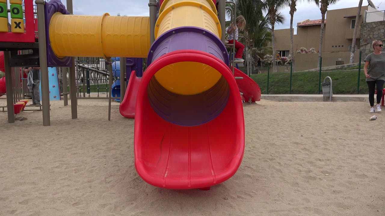 A red slide opening dominates the foreground at a San Jose del Cabo playground, with yellow and purple tube sections above, sand below, and an adult woman standing near the right edge.