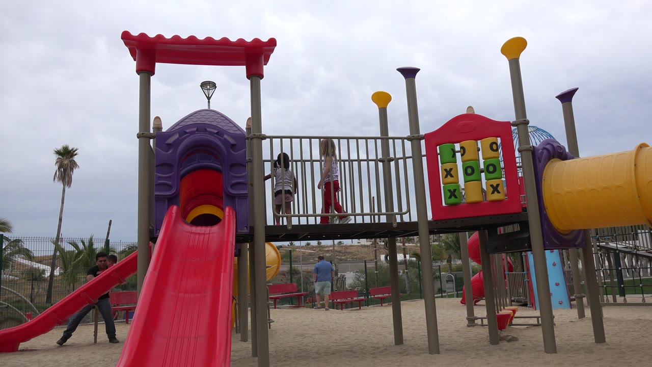 The two girls are still on the raised walkway beside the red slide, with the colorful tic-tac-toe panel and yellow tube slide visible on the playground structure.