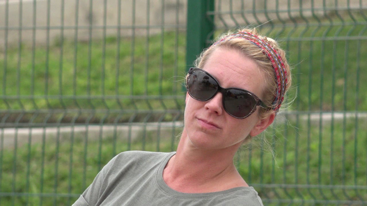 Blonde woman in sunglasses leaning slightly in front of a green fence.