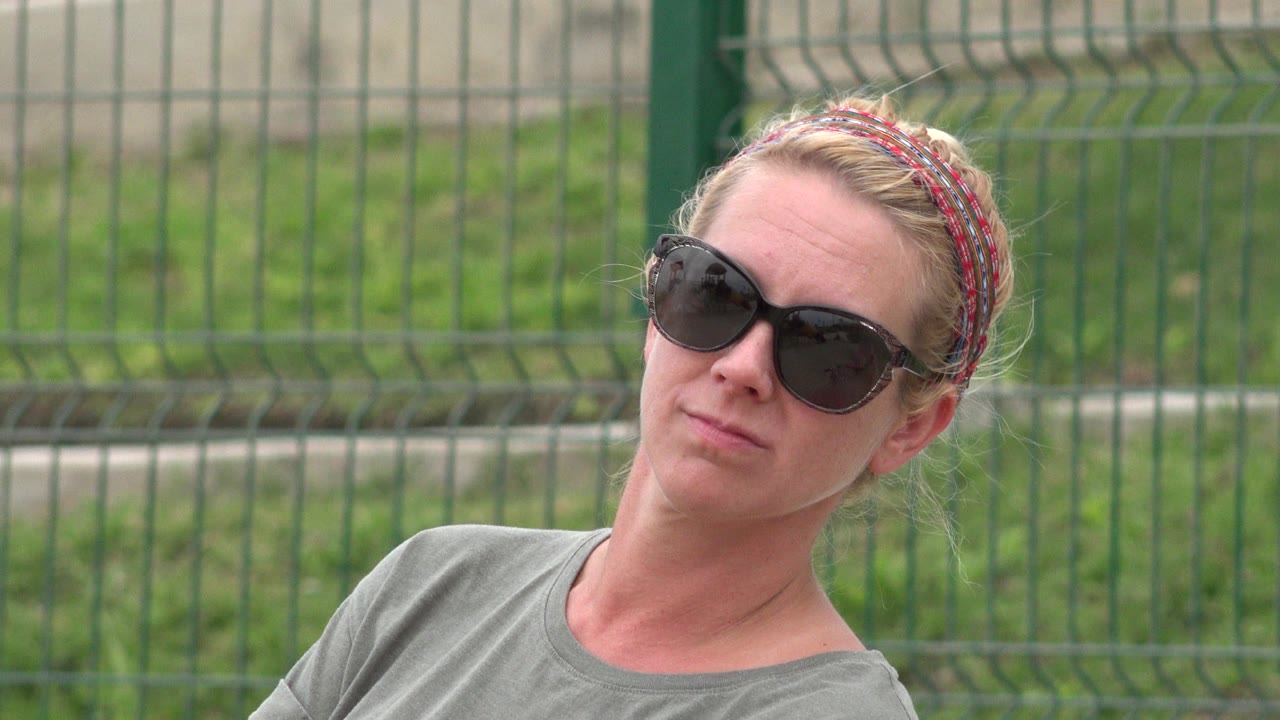 At 0:01, the woman remains close in frame with her head angled, black sunglasses prominent, and the green fence soft behind her in San Jose Del Cabo, Mexico.
