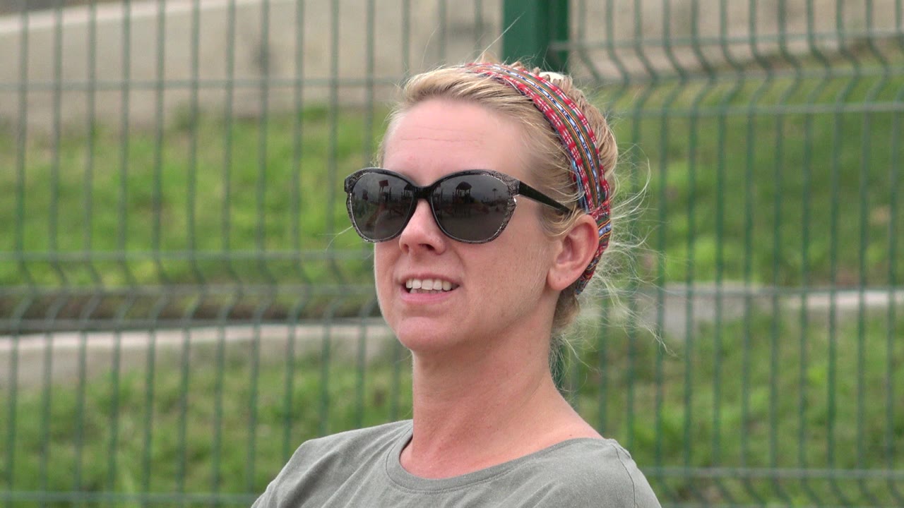 At 0:00, a blonde woman in black sunglasses and a patterned headband smiles in a San Jose del Cabo park with a green fence behind her.