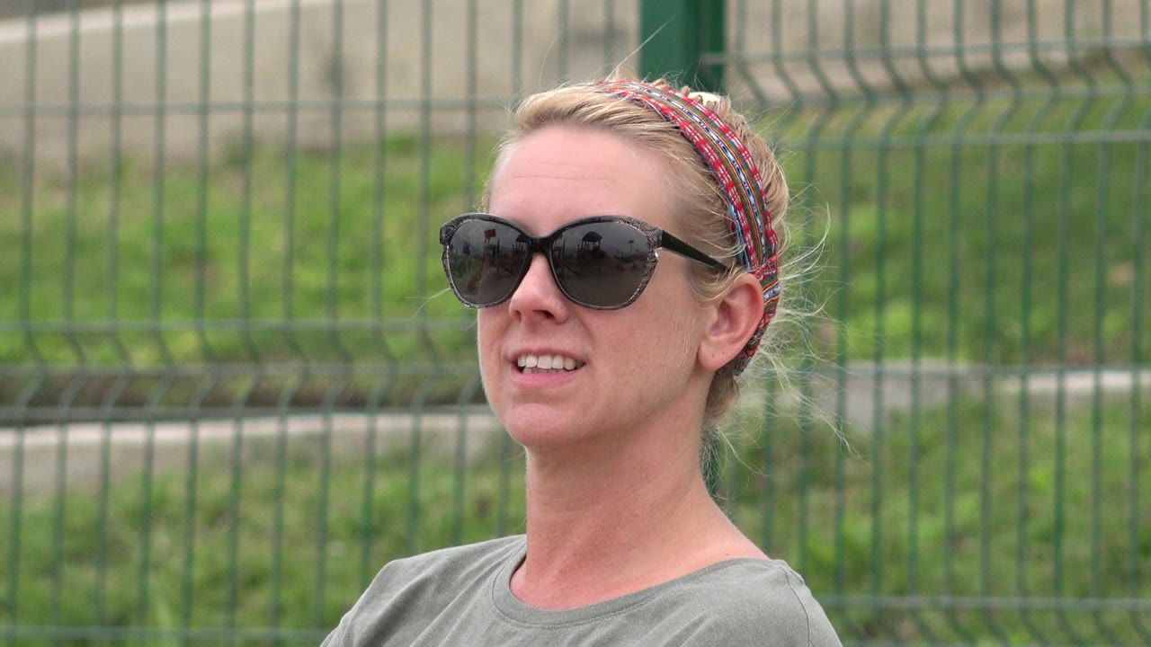 At 0:01, the woman remains in tight profile-leaning view, wearing dark sunglasses with the green metal fence and grass blurred behind her in San Jose del Cabo.