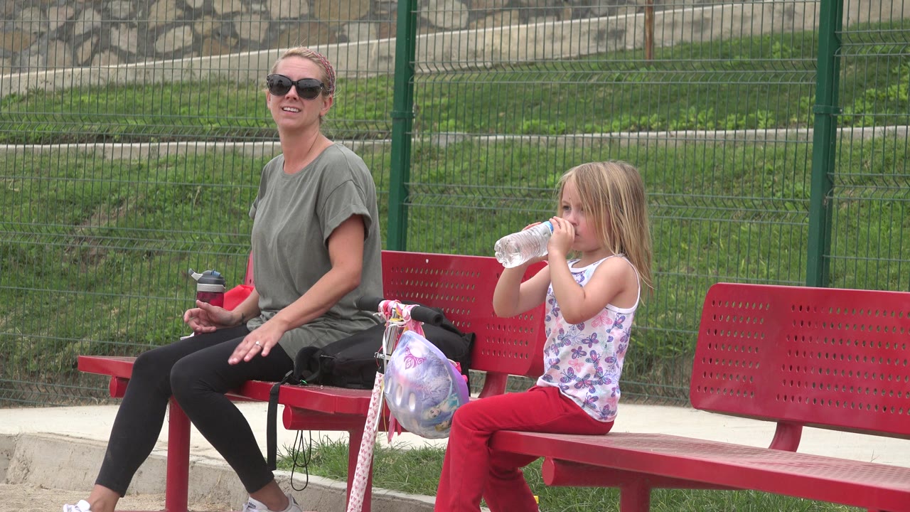 The woman remains seated on the red bench while the young girl drinks from a clear bottle, with the scooter and bags visible between them in San Jose del Cabo.