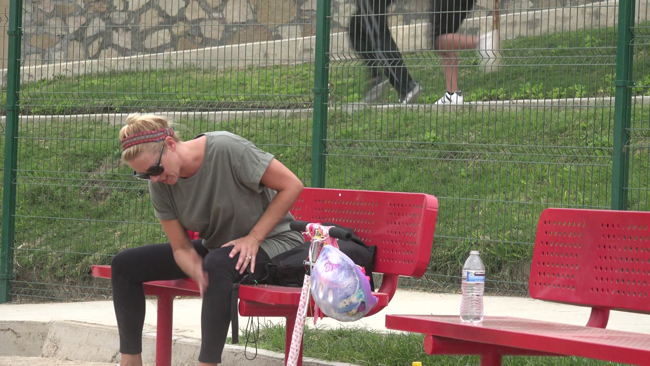 In San Jose Del Cabo, Mexico, a woman in sunglasses leans forward on a red bench with a patterned bag at her side and a water bottle on the pavement nearby.