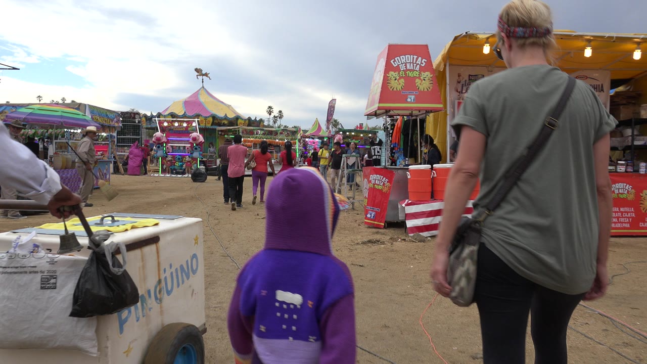 A woman and a child in a purple hood walk away from the camera through fairgrounds in San Jose del Cabo, Mexico, with a pushcart, food stall, rides, and other people visible.