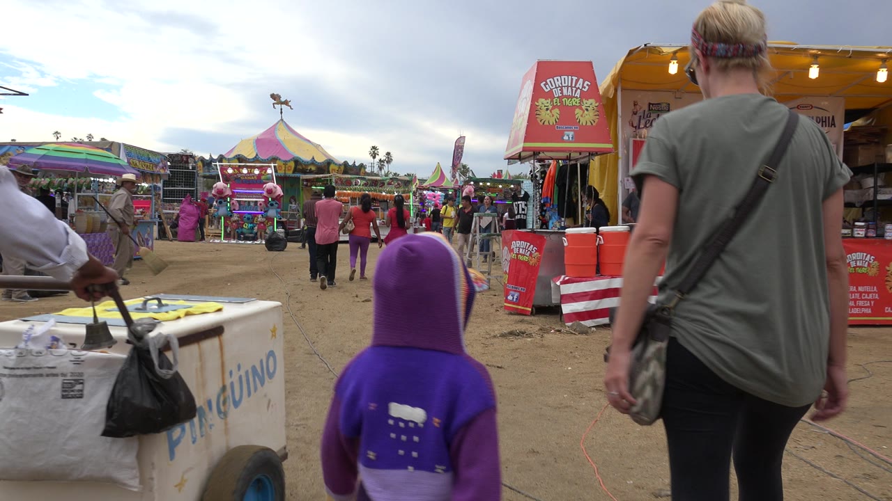 The child in a purple hood remains centered low in frame as an adult walks ahead near vendor carts, colorful fair rides, and pedestrians in San Jose del Cabo, Mexico.