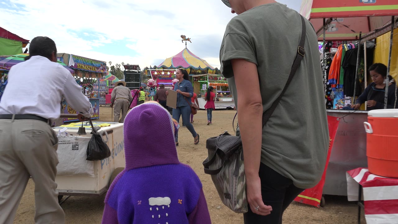 Child in a purple hood and adults walking near amusement park stalls.