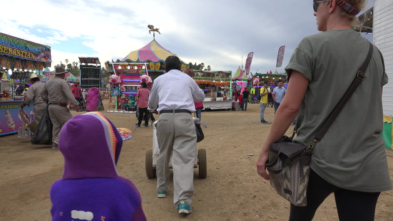 At a San Jose del Cabo carnival, a child in a purple hooded top stands in the foreground while adults walk toward colorful booths and a striped tent.