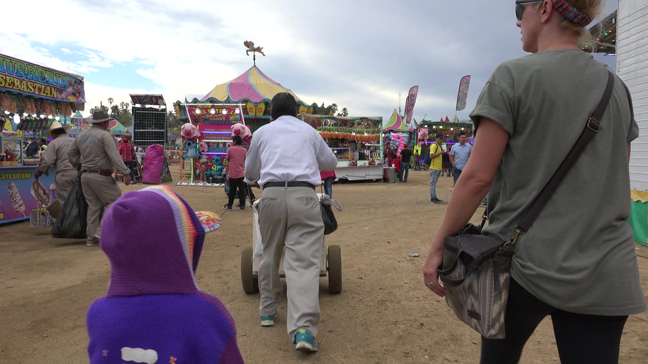 The child in purple remains low in the frame as the adult woman stands at right and a man with a walker moves toward game booths in San Jose del Cabo.