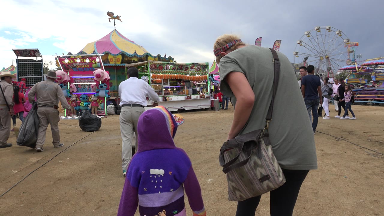 The child and adult remain in the foreground while fair workers stand near a game stall, with a Ferris wheel and colorful canopy behind them in San Jose Del Cabo, Mexico.
