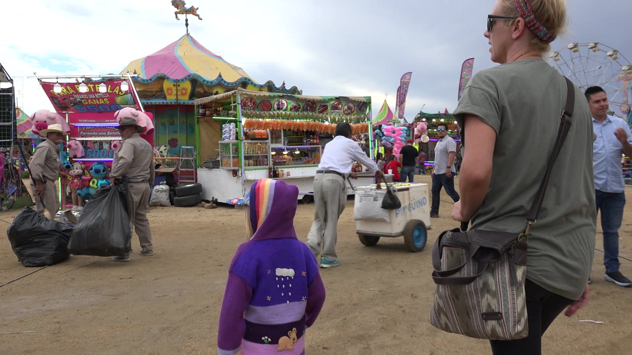 The child remains in the foreground while the adult stands to the right, facing food and game stalls with workers carrying large trash bags in San Jose del Cabo, Mexico.