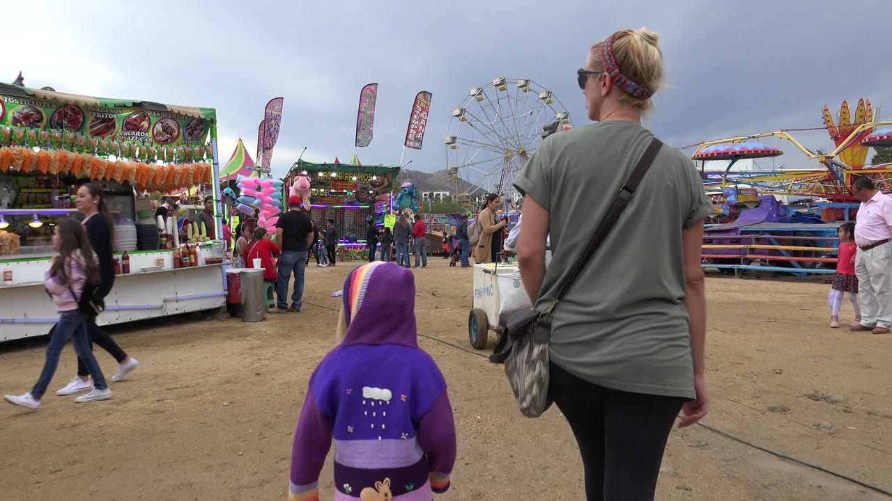 The child in a purple hooded jacket and the woman continue forward at the San Jose del Cabo festival while people cross near bright booths and a Ferris wheel.