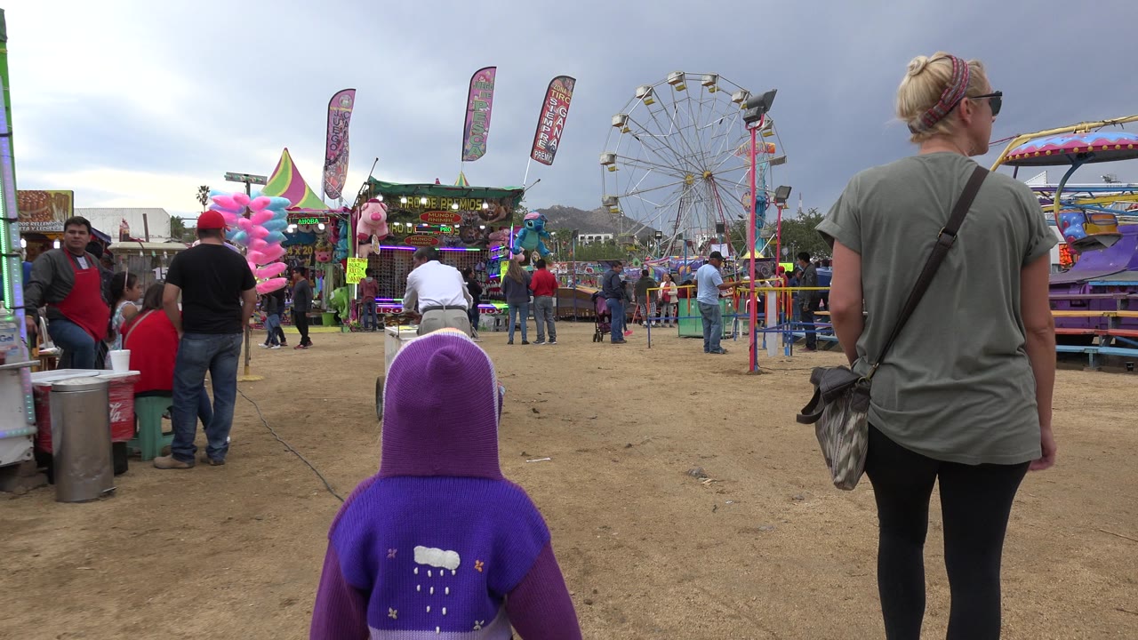 Woman and child walking toward a fairground with rides and vendor stalls.