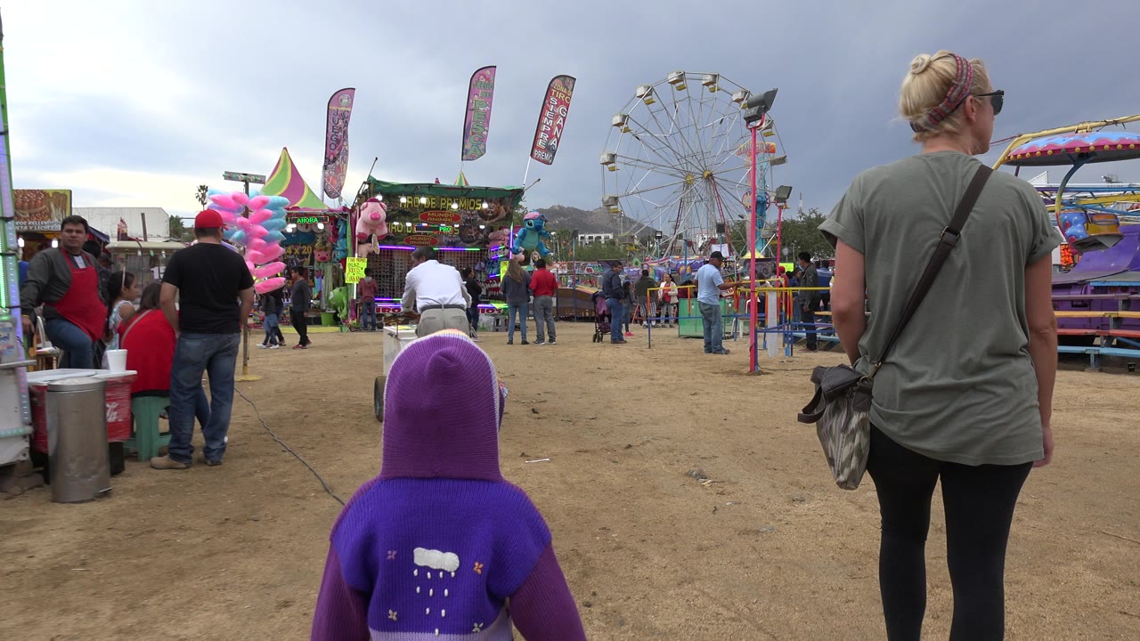 The child in a purple hoodie remains in the lower foreground while the woman walks ahead beside bright ride platforms, vendor lights, flags, and a ferris wheel in San Jose del Cabo, Mexico.