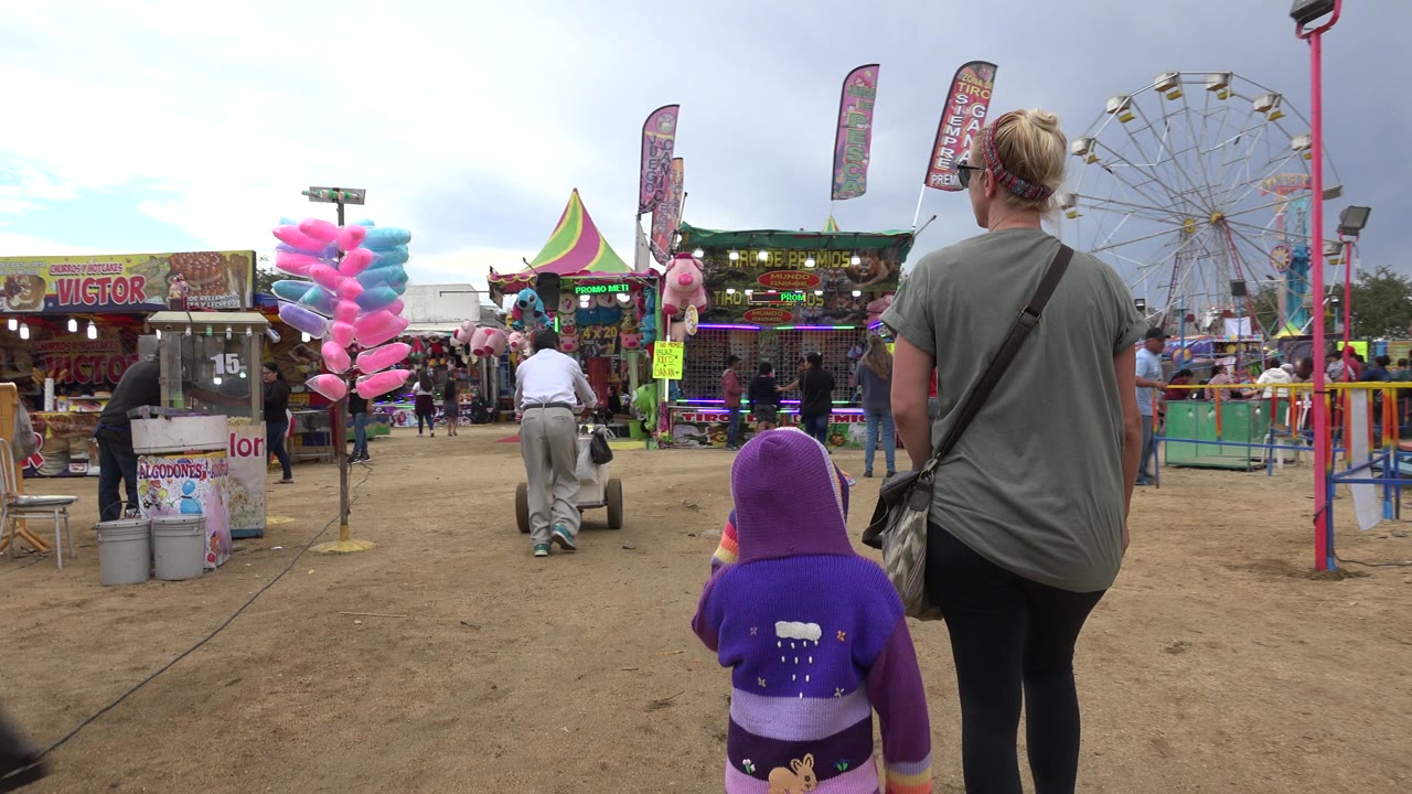 The pair continues along the dirt walkway as colorful cotton candy, lit game booths, stuffed prizes, and the ferris wheel fill the amusement park background in San Jose del Cabo, Mexico.