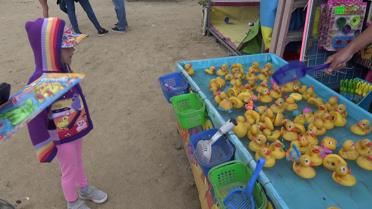 An adult hand reaches toward the blue duck pond game in San Jose Del Cabo, Mexico, with the young girl watching from the left and rows of yellow ducks floating in the play area.