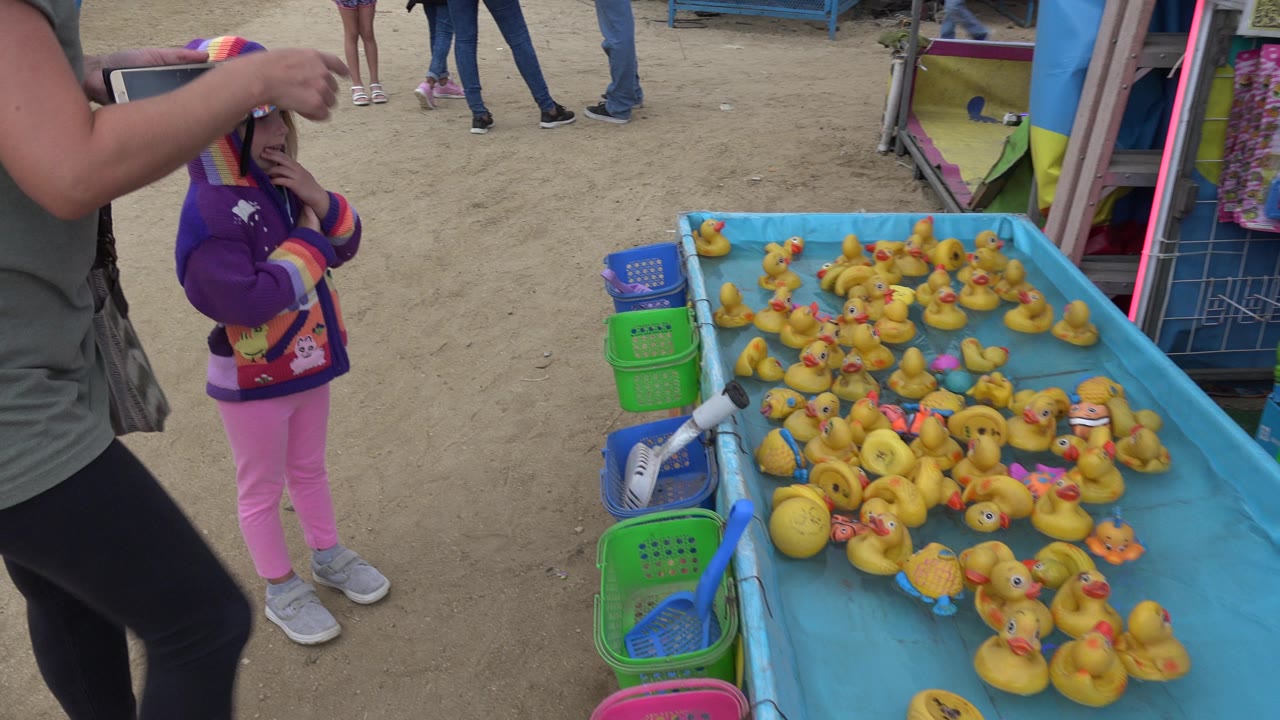 At 0:01 in San Jose del Cabo, Mexico, an adult points across the rubber duck game table while the child stands nearby and fairgoers pass in the background.