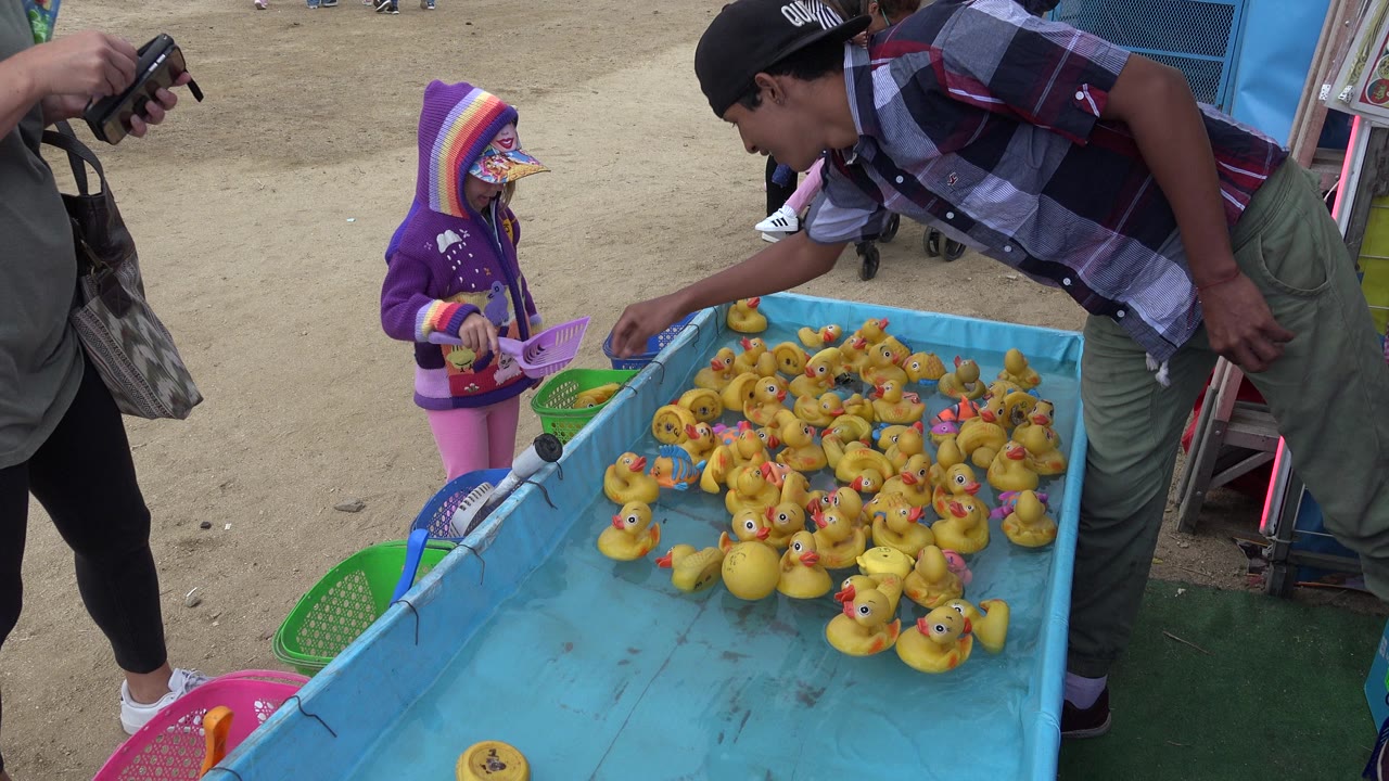 A young girl in a purple hoodie stands at a San Jose del Cabo fair duck fishing game while a booth worker reaches over a blue tank filled with yellow rubber ducks.