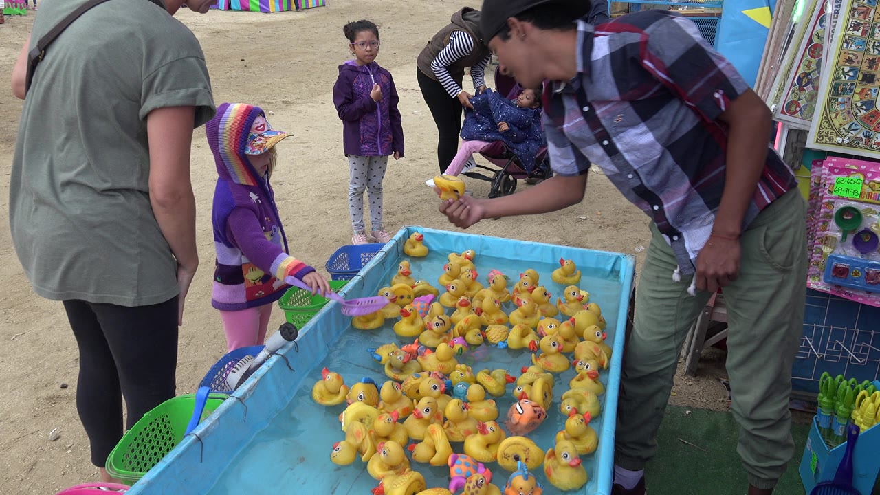 At 0:01 in San Jose del Cabo, Mexico, the child holds a pink net beside a cluster of yellow rubber ducks as an adult leans over the edge of the game.