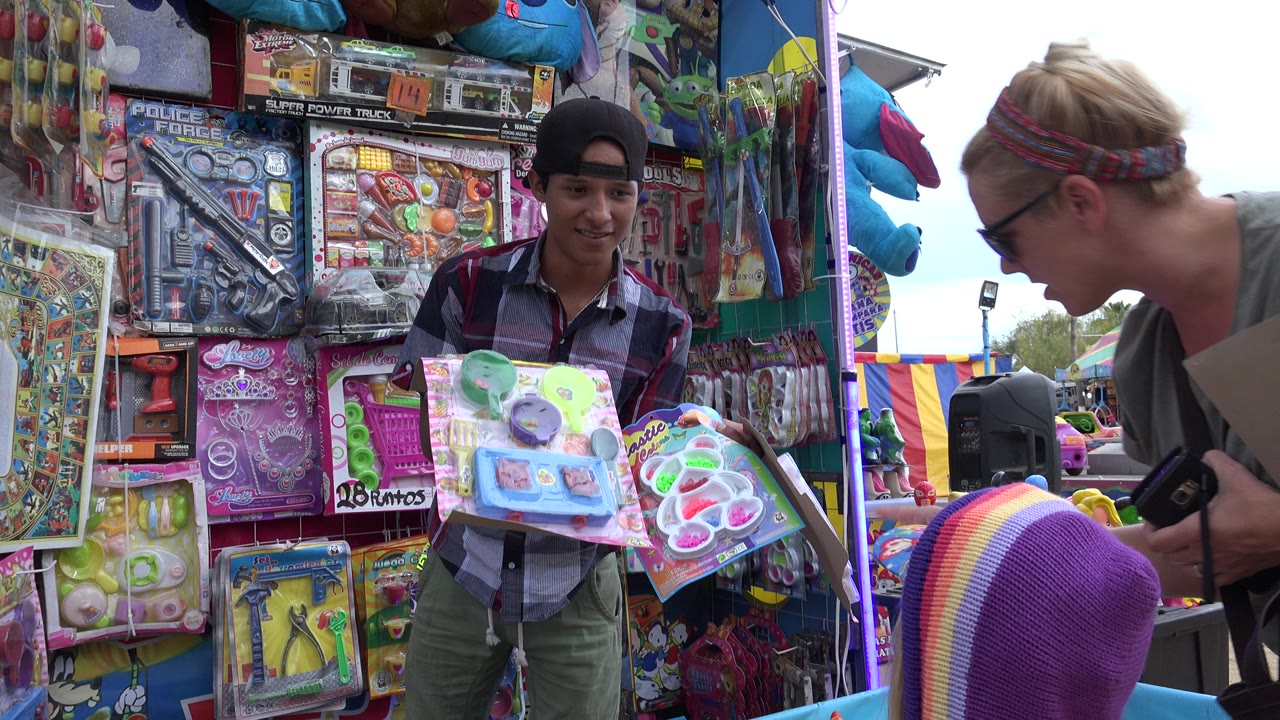 The booth attendant in San Jose del Cabo presents toy packages at the counter while the woman bends toward the child near the floating ducks.