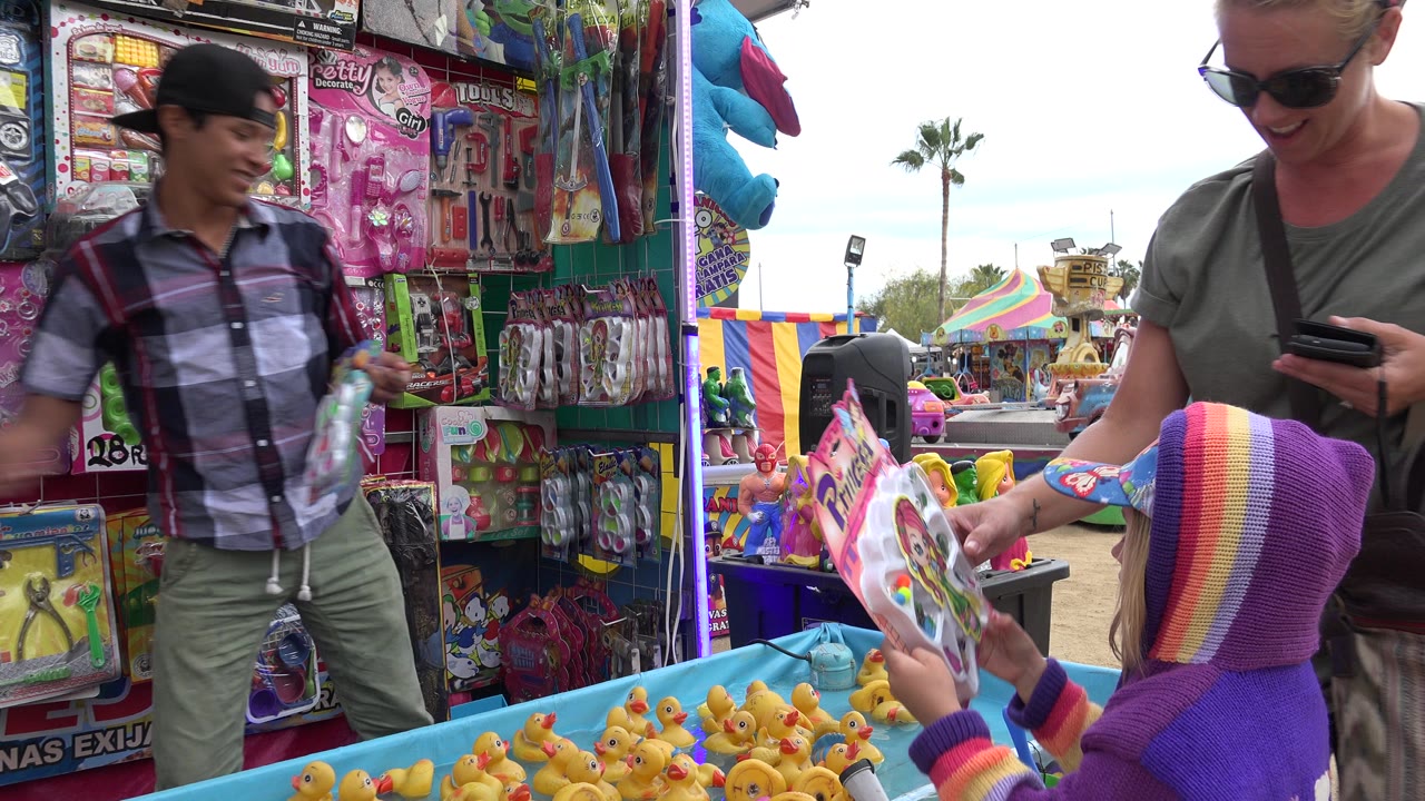 In San Jose del Cabo, Mexico, a carnival worker stands behind a toy-filled booth while a child in a purple rainbow hood reaches near a blue tub of yellow rubber ducks.