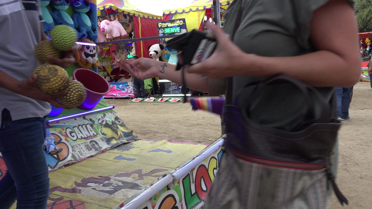 At 0:01 in San Jose Del Cabo, Mexico, the man and woman handle the melons near a game or vendor booth with bright painted panels and hanging prizes behind them.