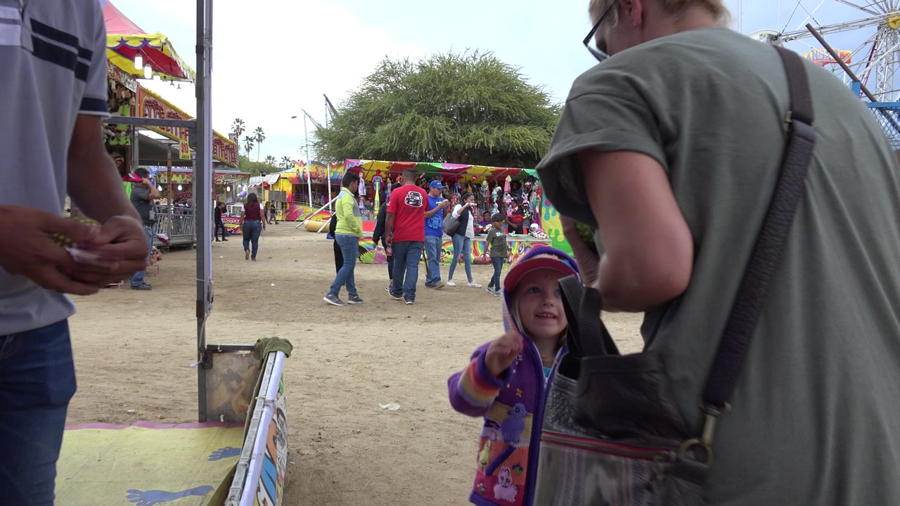The child points from beside the game barrier while adults stand close to the camera and a busy amusement booth area fills the background in San Jose del Cabo, Mexico.