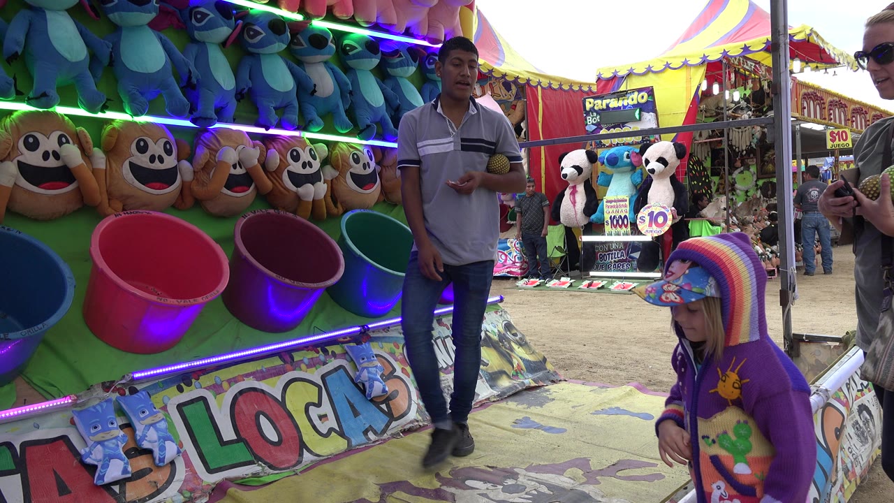 Carnival bucket toss booth with plush prizes, an attendant, and a child in a purple hooded jacket.