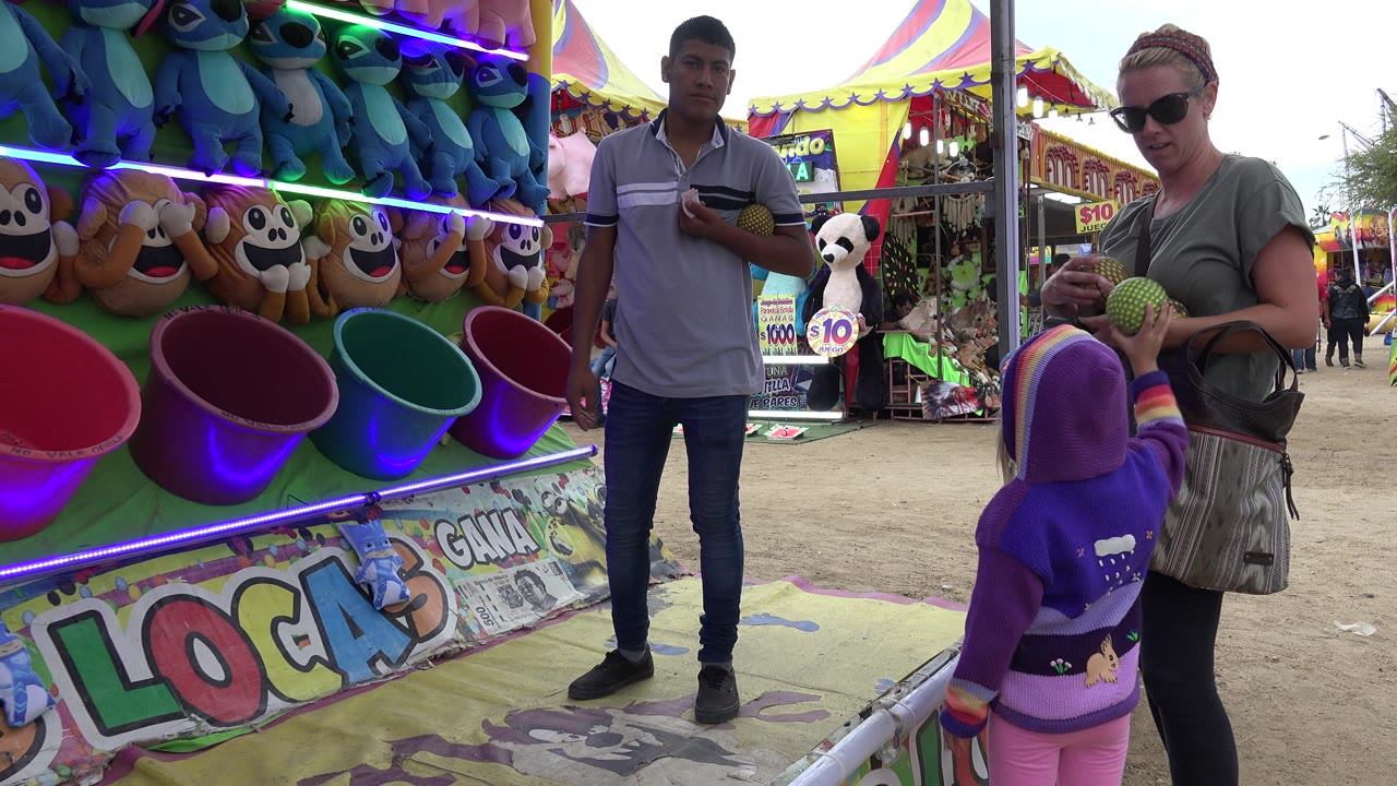 At a San Jose del Cabo carnival-style game booth, a child in a purple hoodie faces bucket targets while a woman holds balls and a male attendant stands nearby.