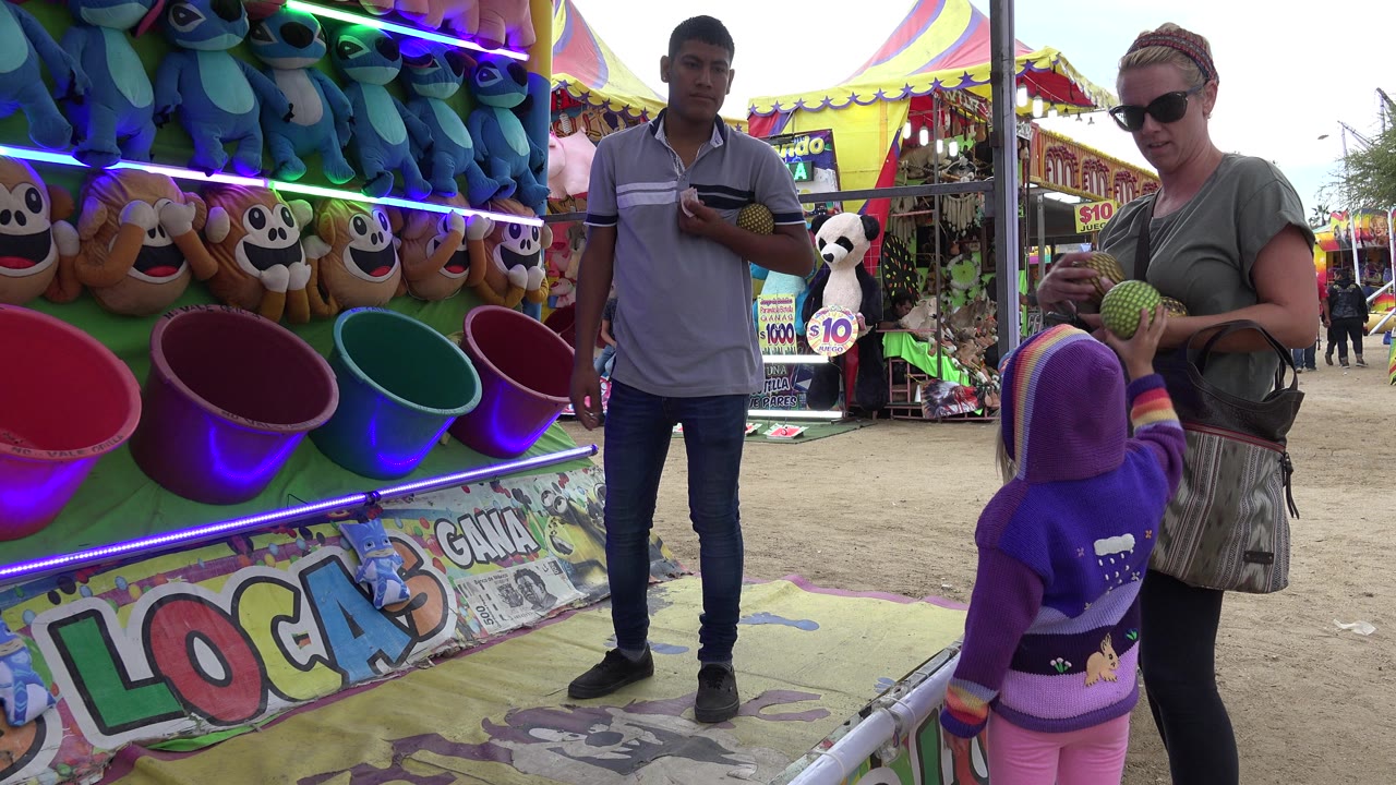 In San Jose del Cabo, the woman stands beside the child with balls in hand as the attendant watches near the colorful bucket targets and hanging plush prizes.