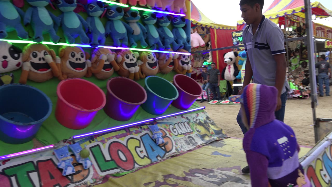 The child remains close to the game rail while a row of red, blue, green, and purple buckets sits below stuffed toys at the San Jose Del Cabo fair booth.