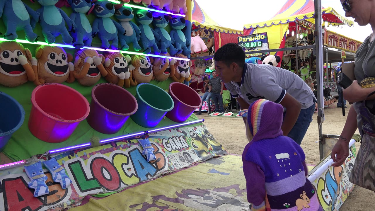 In San Jose del Cabo, Mexico, a child in a purple hooded jacket stands at a colorful ball toss booth while an attendant leans over the counter and plush prizes hang above.