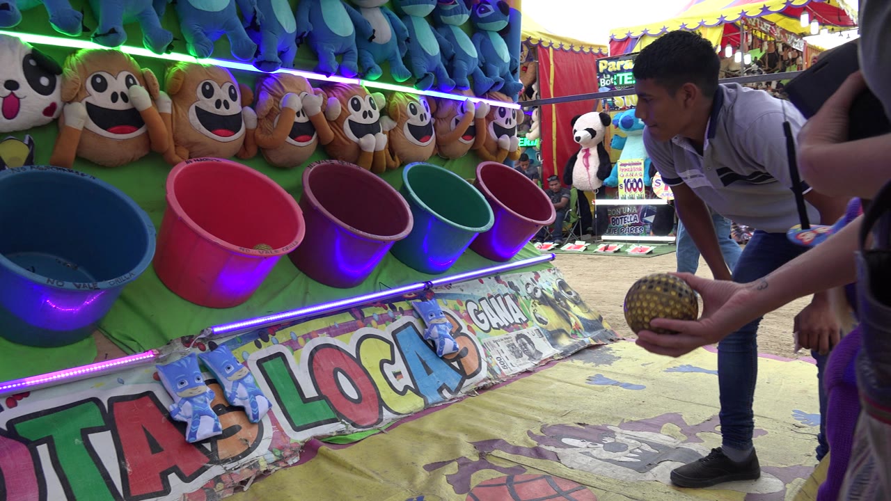 An adult hand holds a patterned ball in front of pink, purple, green, and blue buckets at a San Jose Del Cabo carnival prize game.